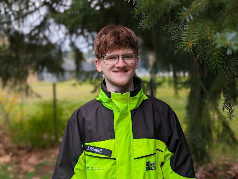 Der Schülerlotse Jannes Schmidt trägt eine gelbe Warnjacke und lächelt in die Kamera.