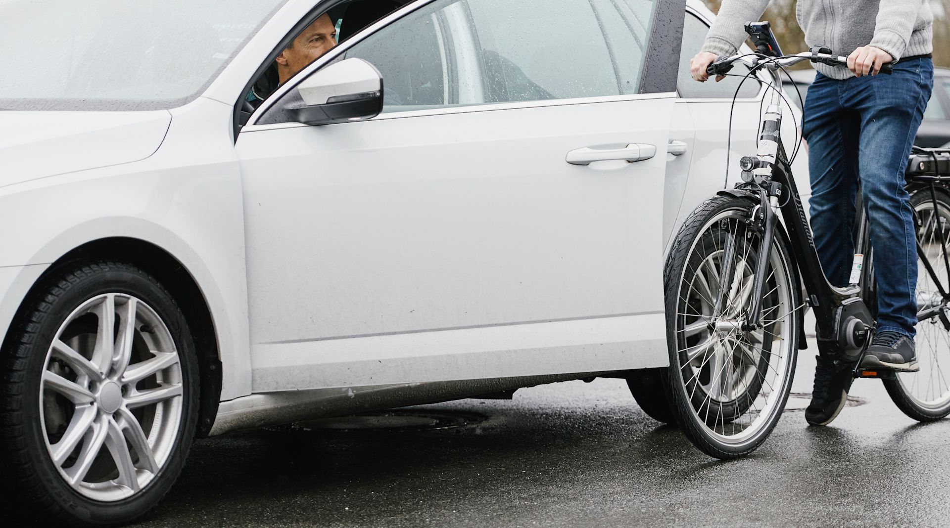 Ein Mann sitzt auf einem Fahrrad, neben ihm ein geparktes Auto, aus dem ein anderer Mann gerade aussteigen möchte. Die Autotür ist geöffnet.