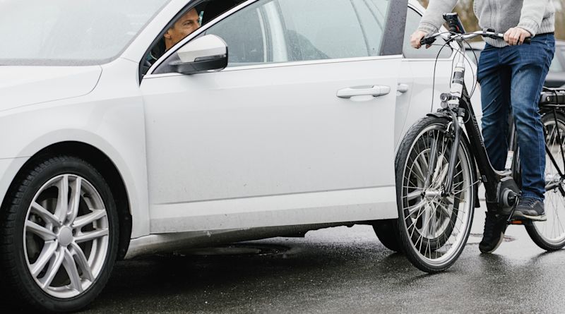 Ein Mann sitzt auf einem Fahrrad, neben ihm ein geparktes Auto, aus dem ein anderer Mann gerade aussteigen möchte. Die Autotür ist geöffnet.