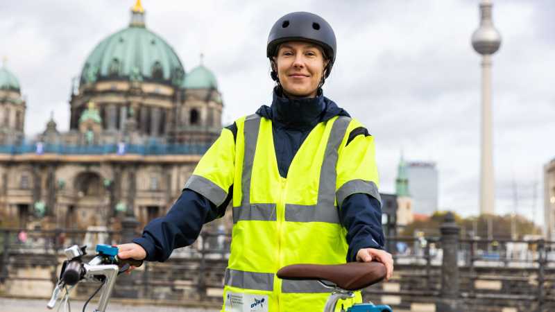 Eine Frau steht mit einem Fahrrad vor dem Berliner Dom. Am Lenker und unter dem Sattel sind technische Geräte montiert. Sie ist mit einer gelben Warnweste und einem Helm bekleidet.