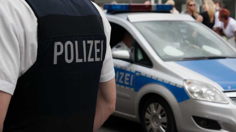 Ein Polizist ist von hinten zu sehen, auf seiner Weste steht in großen Lettern „POLIZEI“. Im Hintergrund ist ein blaues Polizeifahrzeug zu sehen. 
