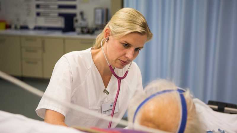 Das Bild zeigt eine Ärztin, die mit einem Stethoskop einen im Bett liegenden Patienten abhorcht.