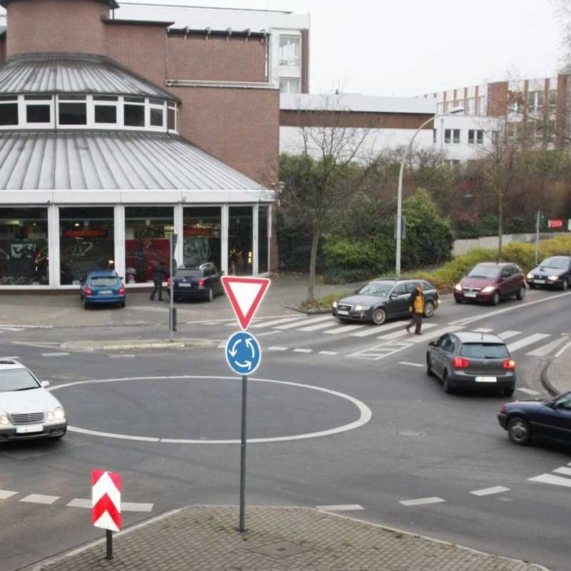 Man sieht einen Kreisverkehr innerhalb einer Ortschaft mit einigen Autos und Fußgängern auf Zebrastreifen.