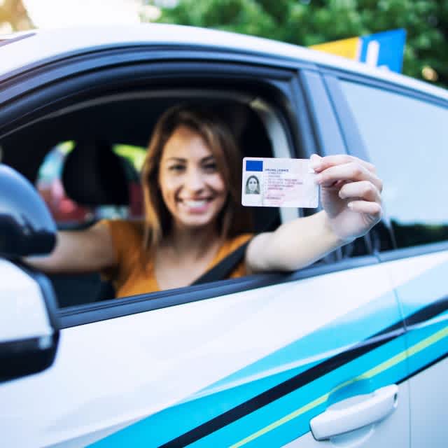 Eine junge Frau sitzt am Steuer eines Pkw und hält lächelnd ihren Führerschein in die Kamera. 
