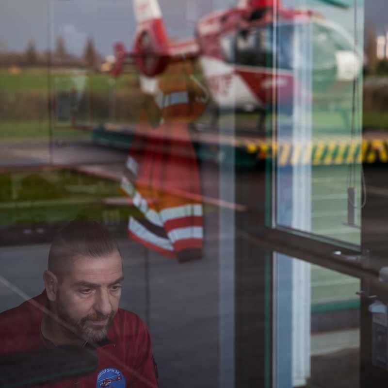 Das Bild zeigt einen Rettungsflieger in der Zentrale vorm Hubschrauber-Landeplatz. Die Kamera blickt durch die leicht spiegelnde Außenscheibe, in welcher das Spiegelbild des Hubschraubers zu sehen ist.