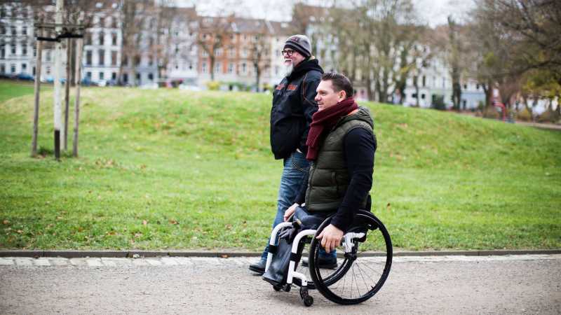 Man sieht zwei Männer von vorne im Park spazieren gehen, einer hat keine Beine und sitzt im Rollstuhl.