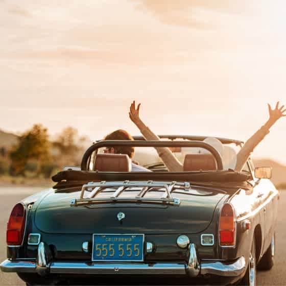 Das Bild zeigt ein Pärchen in einem Oldtimer Cabrio von hinten, die Frau streckt die Arme nach oben und sie fahren in den Sonnenuntergang.