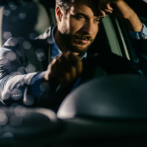 Ein Mann sitzt am Steuer seines Autos und hat seinen Kopf auf seinen Arm gestützt. 
