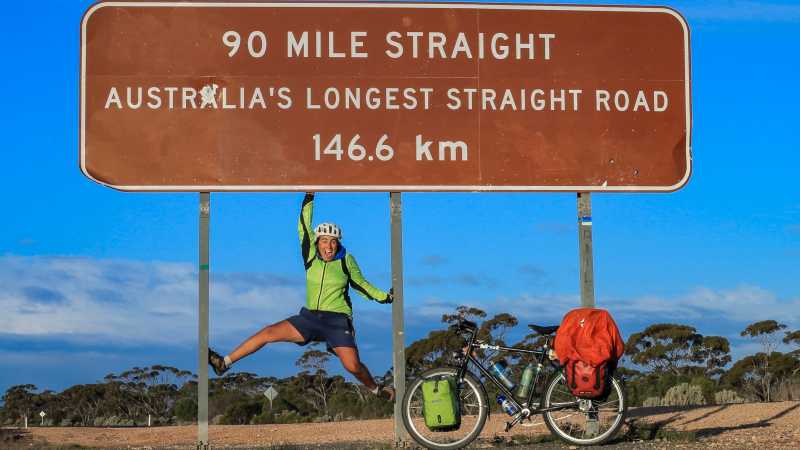 Luisa Rische springt vor einem Schild: 90 Miles straight, Australias longest straight road, 146,6 km. 