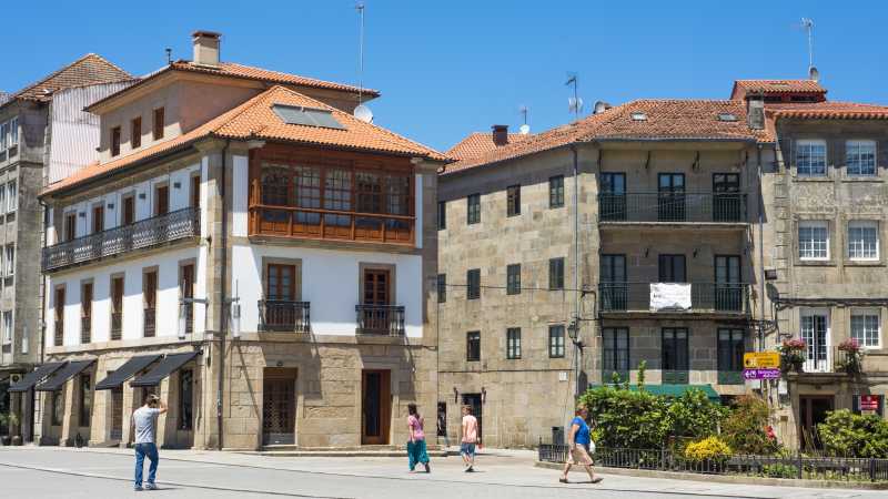 Das Bild zeigt einen Schienenübergang vor einer Häuserfront in der Stadt Pontevedra, vor der einige Fußgänger und Fußgängerinnen langlaufen.