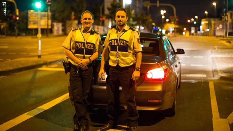 Die Polizisten Rainer Fuchs und Marco R. stehen vor ihrem Einsatzwagen an einer leeren Kreuzung in der Nacht und lächeln.
