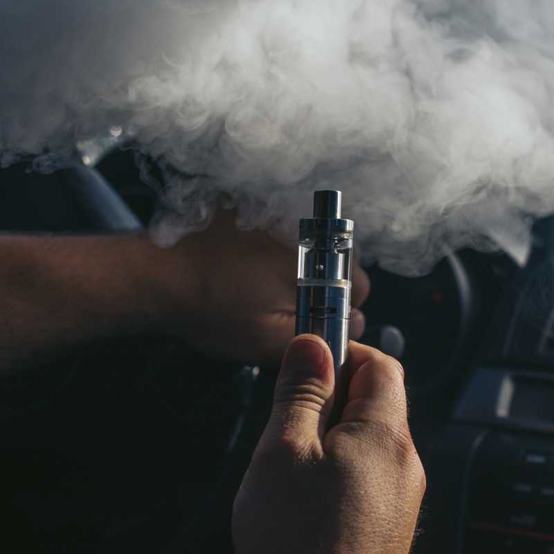 Eine Hand hält im Auto eine E-Zigarette. Darüber ist eine eine dichte Rauchwolke zu sehen