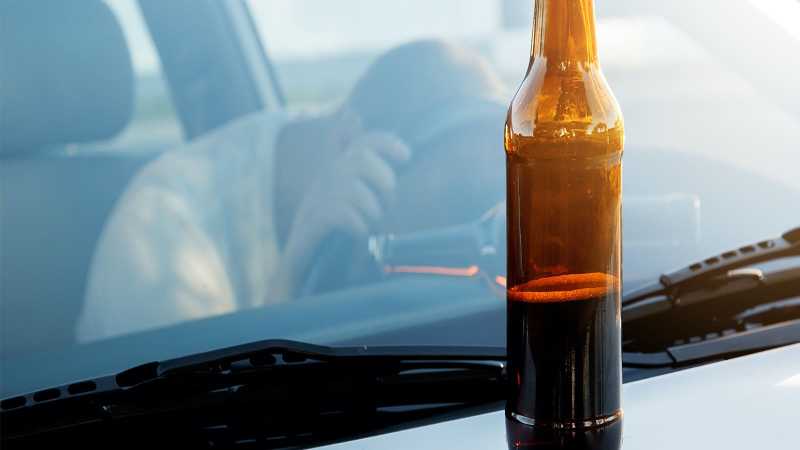 Das Bild zeigt eine halb leer getrunkene Flasche Bier auf einer Motorhaube. Im Hintergrund sieht man durch die Frontscheibe des Autos einen jungen Fahrer, der verzweifelt den Kopf auf dem Lenkrad auflegt.