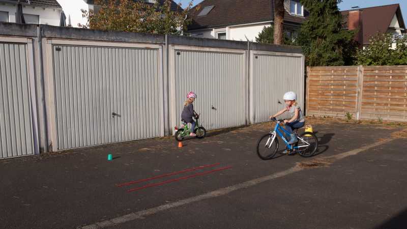 zwei Kinder fahren auf ihren Fahrrädern in einem Garagenhof durch eine aus zwei Seilen gelegte Spur.