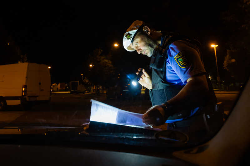 Ein Polizist überprüft bei Dunkelheit mit einer Taschenlampe in der Hand Unterlagen. 