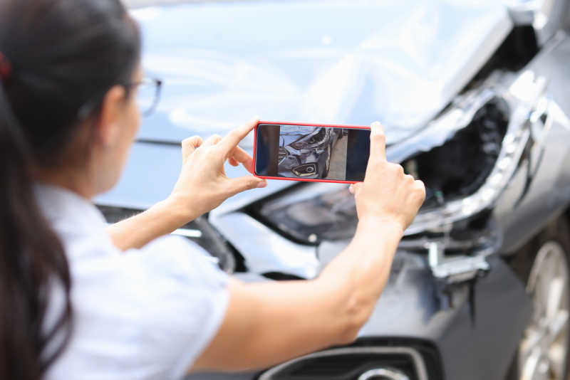 Eine Frau fotografiert einen Unfallschaden an der Motorhaube eines grauen Pkw.