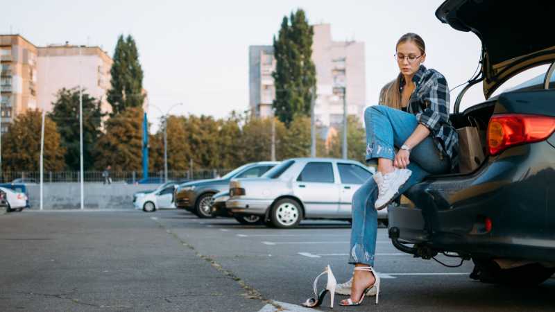 Eine Frau sitzt im Kofferraum eines Wagens, um die Schuhe zu wechseln. Ihre hohen Absätze tauscht sie in bequeme Sneaker ein. Das ist deutlich verkehrssicherer.