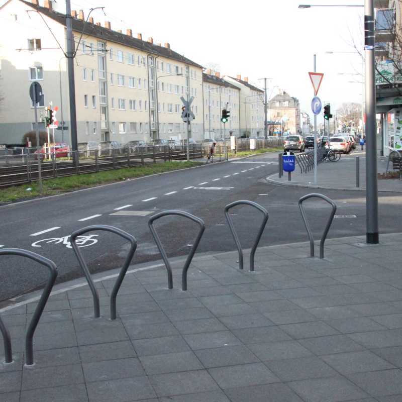 Abstellbügel für Fahrräder stehen auf dem Gehweg. Im Hintergrund ist eine Straße mit Fahrradschutzstreifen zu sehen. 