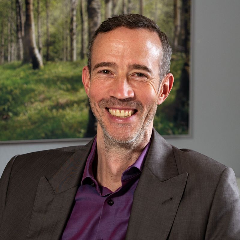 Vor einem unscharfen Waldbild sitzt ein Mann im Anzug mit lila Hemd. Es handelt sich um ein Porträt von Christian Müller, Verkehrspsychologe.