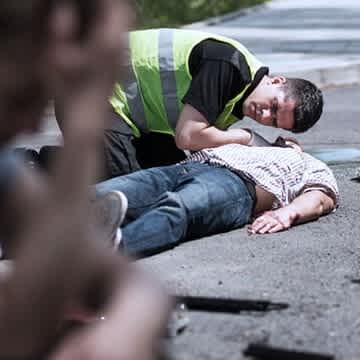 Man sieht einen Unfallort, auf dem Fahrzeugteile liegen, ein Junge sitzt auf dem Boden und hält sich den Kopf, während ein Mann sich über einen weiteren Jungen beugt und dessen Atem prüft.