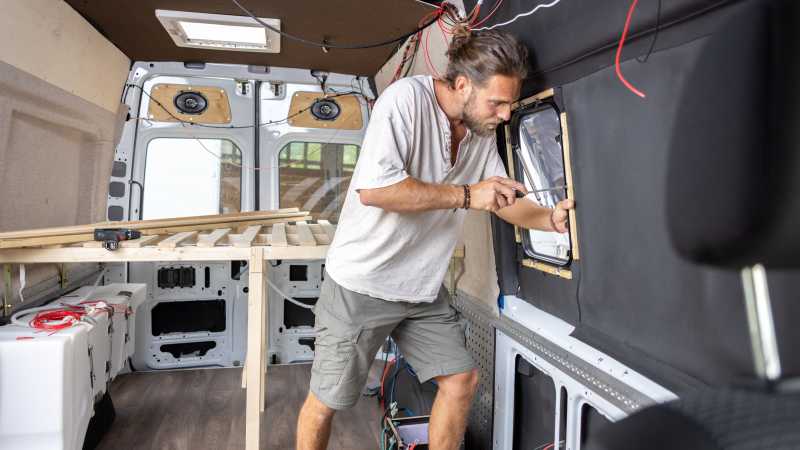 Ein junger Mann setzt in seinen selbst ausgebauten Camper-Van ein neues Fenster ein.