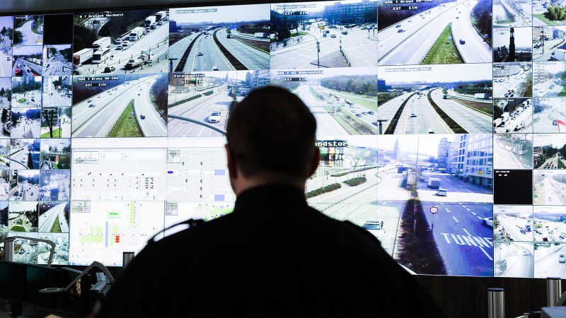 Man sieht einen Mann vor einer großen Leinwand sitzen, die Überwachungsbilder der Straßen Hamburgs zeigt.