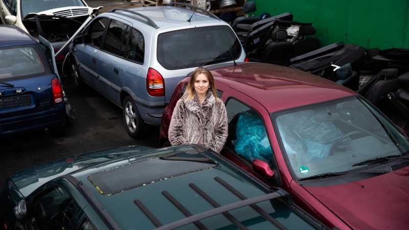 Das Bild zeigt eine junge Frau aus der Vogelperspektive, die zwischen mehreren Autos auf einem Schrottplatz steht.