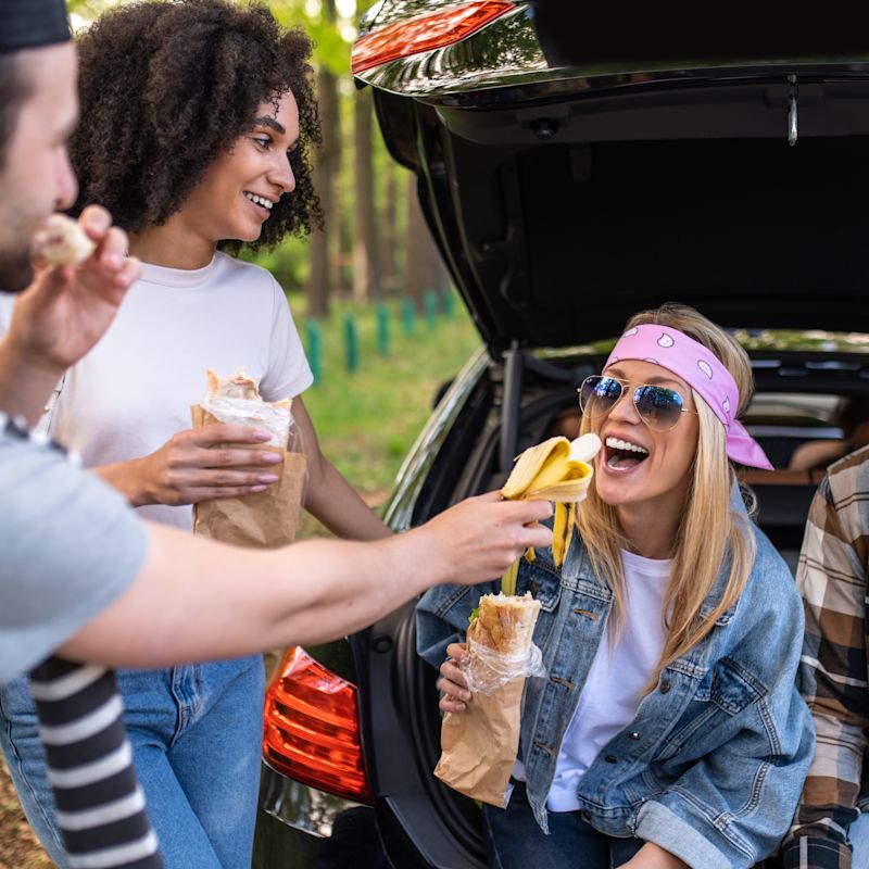 Eine Gruppe von vier jungen Menschen sitzen in und stehen vor einem offenen Kofferraum und machen eine Pause auf dem Weg zum Festival. Sie essen Brote und eine Banane.