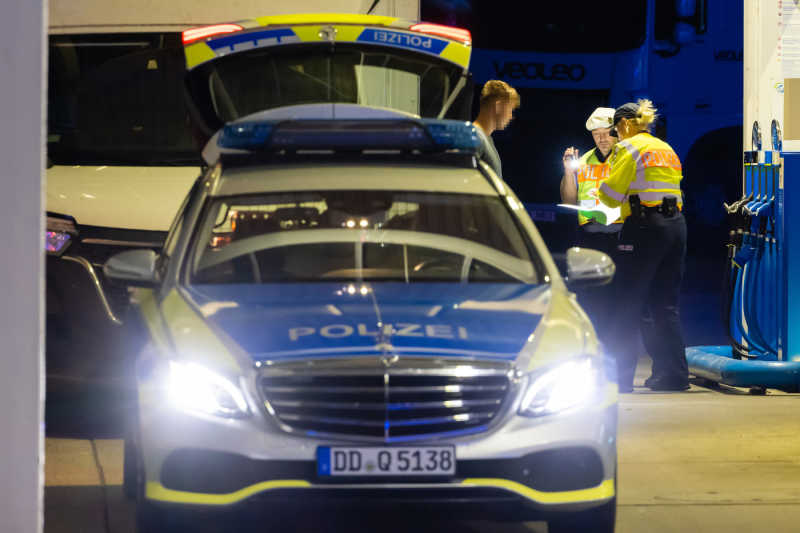 Zwei Polizisten kontrollieren die Unterlagen eines jungen Mannes. Er ist Lkw-Fahrer. Im Vordergrund steht ein Polizeiwagen mit eingeschaltetem Licht.
