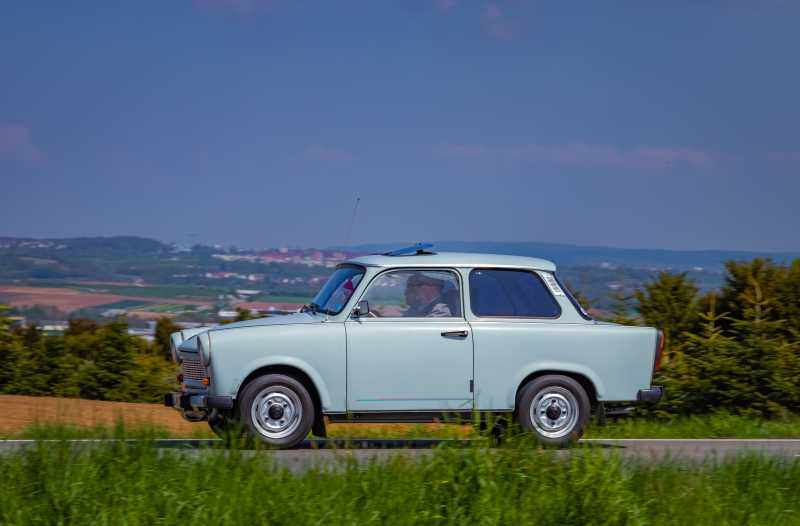 Das Bild zeigt ein altes Auto aus den 70ern auf einer Landstraße mit einer ländlichen Landschaft im Hintergrund.