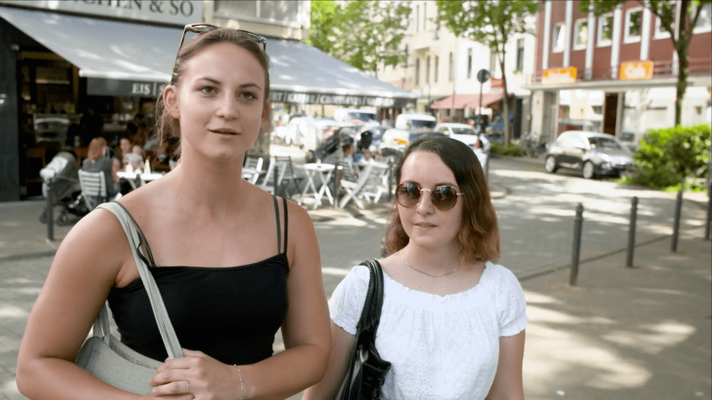 Das Bild zeigt zwei junge Frauen in einer Fußgängerzone, die interviewt werden.
