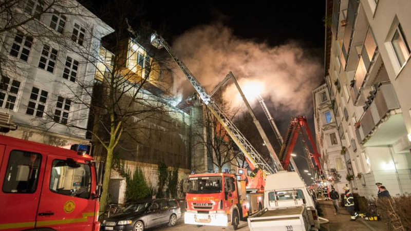 Das Bild zeigt ein brennendes Gebäude, aus dem viele Rauch empor steigt. In der engen Straße stehen mehre Feuerwehrfahrzeuge, die versuchen das Feuer zu löschen. 