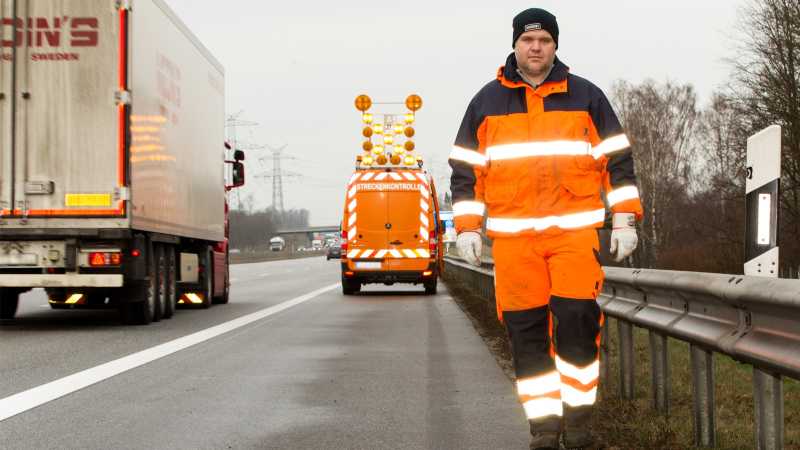 Der Straßenwärter André Schlüter läuft den Standstreifen entlang, neben ihm fährt ein LKW und im Hintergrund sieht man das Fahrzeug der Streckenkontrolle.