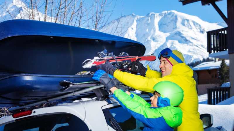 Ein Mann mit Kind belädt die Dachbox seines Autos mit Wintersport-Equipment. 