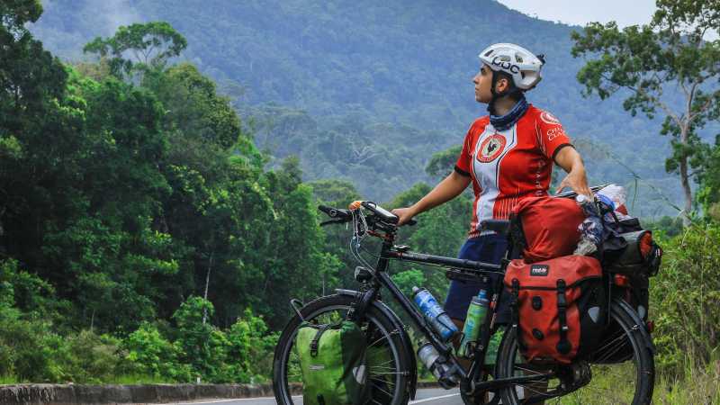 Luisa Rische steht mit ihrem Fahrrad an der Seite einer Straße mit einem tropischen Regenwald im Hintergrund.
