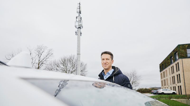 Portrait von Prof. Christoph Degen. Er steht neben seinem Auto auf einem Parkplatz, hinter ihm ist ein Mobilfunkmast zu sehen.