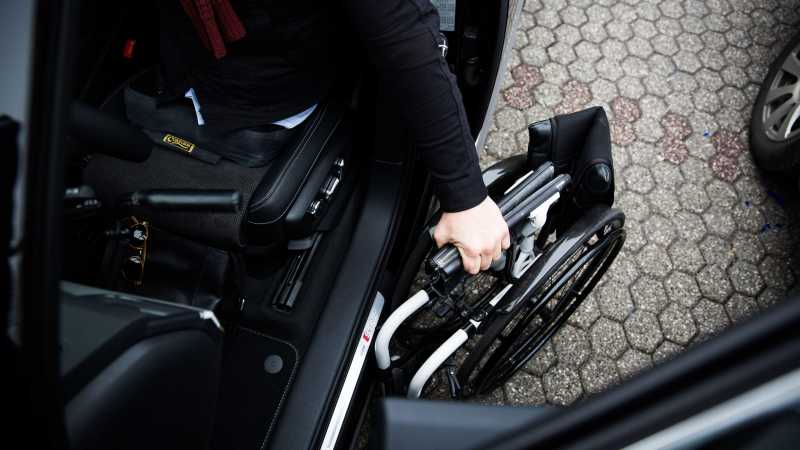 Man sieht einen Mann auf dem Fahrersitz eines Autos, der seinen zusammengeklappten Rollstuhl in der Hand hält.