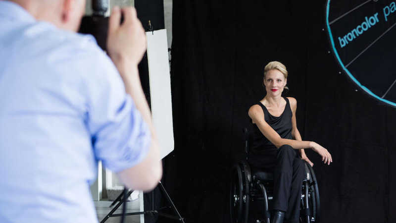 Nina Wortmann sitzt in schwarz gekleidet vor einem schwarzen Hintergrund in ihrem Rollstuhl und wird fotografiert.