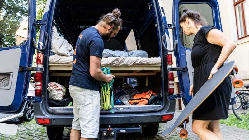 Eine Frau steht mit einem Skateboard in der Hand vor dem offenen Kofferraum eines Vans. Daneben steht ein Mann. Er hat einen Campingstuhl in der Hand. 