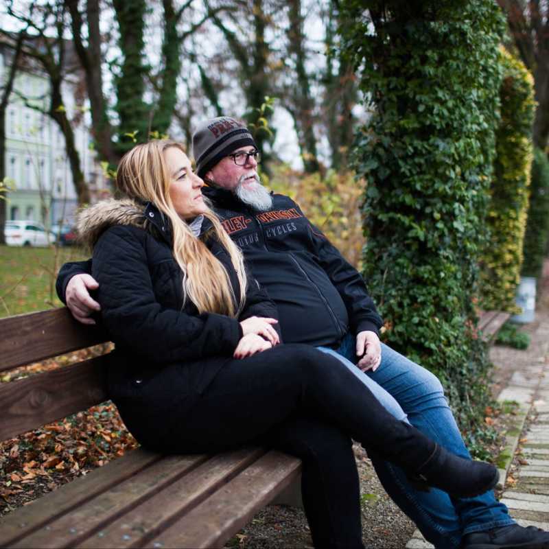 ein Mann und eine Frau sitzen auf einer Bank in einem Park, er hat den Arm um sie gelegt.