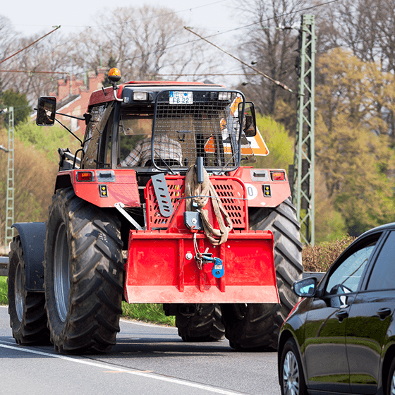 Ein roter Traktor fährt eine Landstraße entlang, hinter ihm fährt ein weiteres Auto.