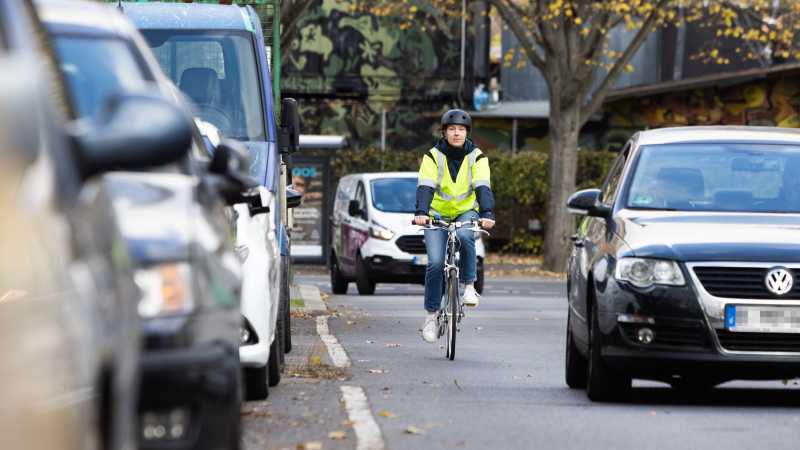 Eine Frau fährt mit dem Fahrrad auf einer Straße. Sie wird von einem dunklen Pkw überholt.