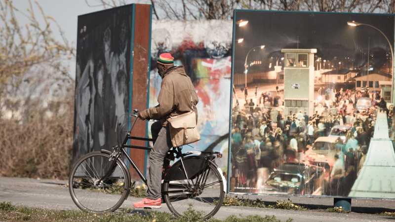 Das Bild zeigt einen Fahrradfahrer, der an einem als Denkmal erhaltenen Teil der Mauer vorbeifährt. 