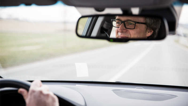 Man sieht die Windschutzscheibe eines Autos von Innen und das Gesicht eines Mannes mit Brille im Rückspiegel.