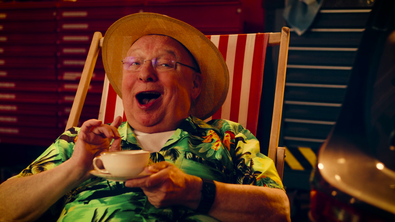 Hassknecht sitzt mit Strohhut und Hawaii-Hemd bekleidet in einem Liegestuhl, hält eine Teetasse und lacht in die Kamera. 