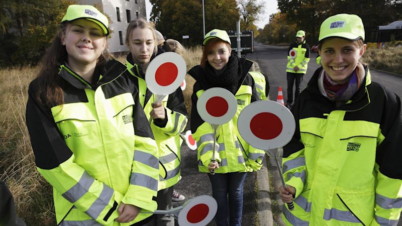 Eine Gruppe von Schülerlotsinnen und Schülerlotsen lächelt in die Kamera. Sie haben gelb reflektierende Jacken an und halten Verkehrskellen in der Hand. 