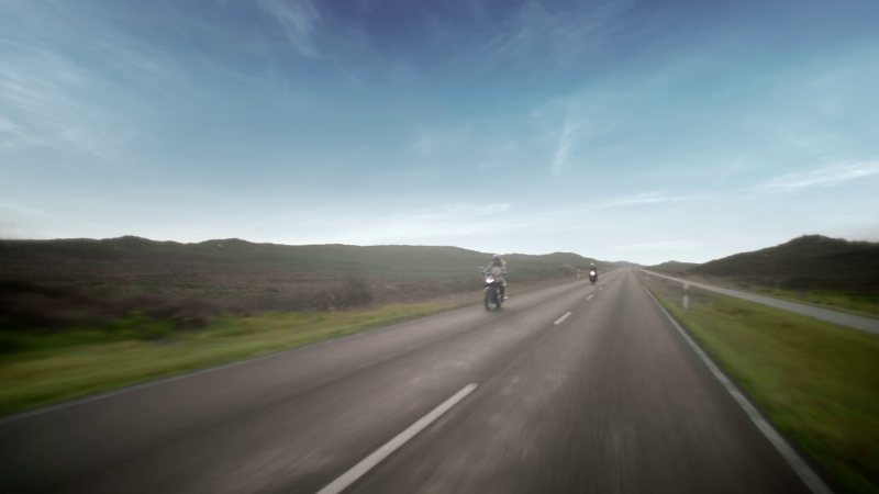Zwei Motorräder sind auf einer Landstraße unterwegs. Der Himmel ist blau, grüne Hügel zeichnen sich am Horizont ab. 