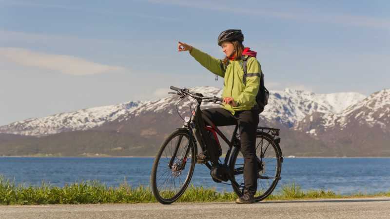 Eine Frau mit Helm hält mit ihrem E-Bike an und zeigt in die Ferne. Im Hintergrund liegen verschneite Berge