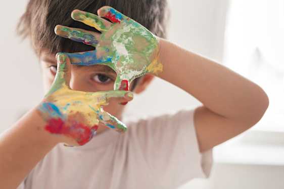 Niño con manos llenas de pintura haciendo una ventana con sus manos por dónde asoma su ojo