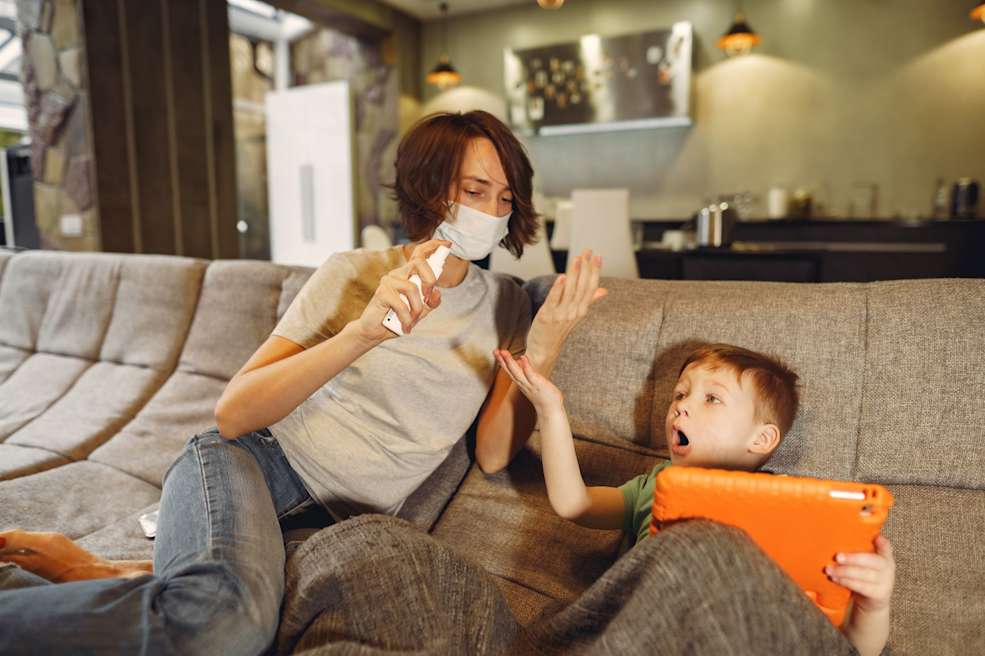 niño y mujer sentados en un sofa ella con mascarilla y alcohol y niño con tablet